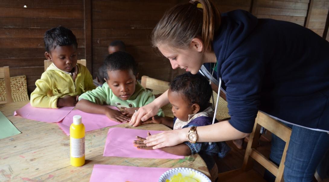 Projects Abroad Kinderopvang vrijwilliger helpt een jong kind met vingerveren tijdens een creatieve activiteit in een dagzorgcentra in Madagaskar. Projects Abroad vrijwilliger werkt met kinderen in Madagaskar en help met handschilderen in een dagzorgcentra.